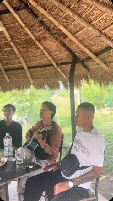 Three musicians under a thatched hut at MEMC Wild Roots Farmhouse, one playing an acoustic guitar.