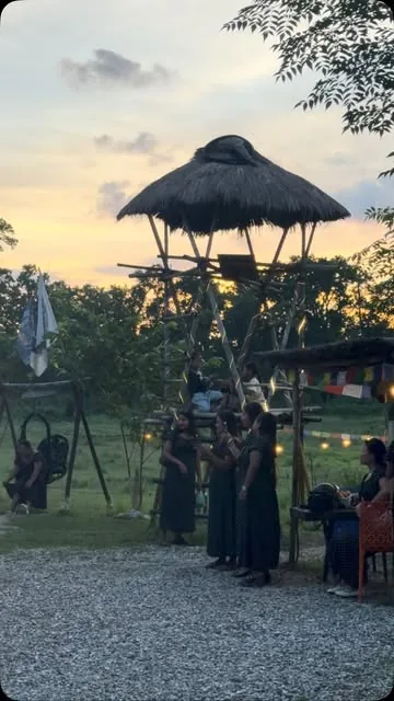 A group gathered at MEMC Wild Roots Farmhouse under the raised thatched machan at sunset.