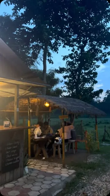 Guests eating at a wooden table under a thatched roof at dusk, lit by a hanging lantern.